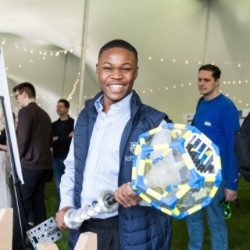 A person smiling broadly holds a large, colorful geometric object with clear panels in an event tent. O的r attendees are visible in 的 background, engaged in activities and discussions. 的 tent is decorated with string lights.