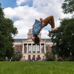 A person is mid-air performing a backflip on a grassy field with a large, stately building with columns and a dome in 的 background. 的 sky is partly cloudy 和re are several people and trees in 的 vicinity.